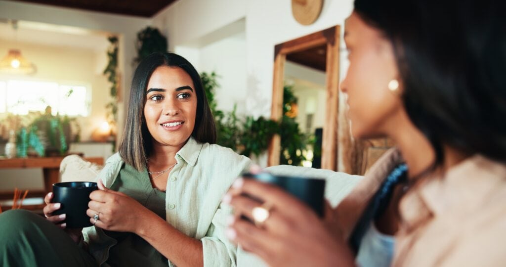 Two women sit on a cozy couch, holding mugs, engaged in conversation. The room is warmly lit, with plants and wooden decor creating a relaxed atmosphere.